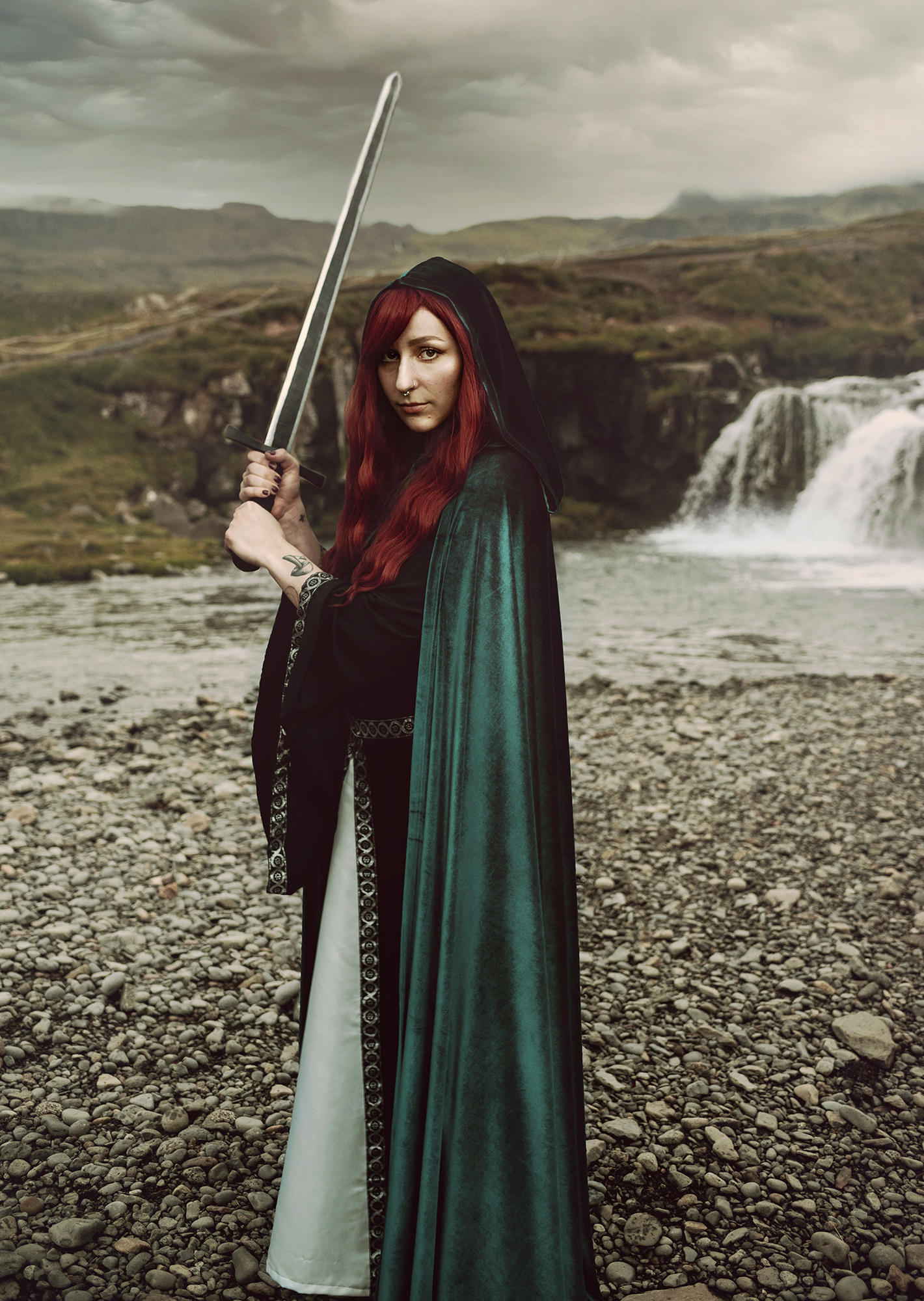 A white woman with red hair holds a sword as she poses in front of a waterfall wearing a black and silver medieval dress and a green velvet cape for a fantasy photo by Kendra Coleen Photography
