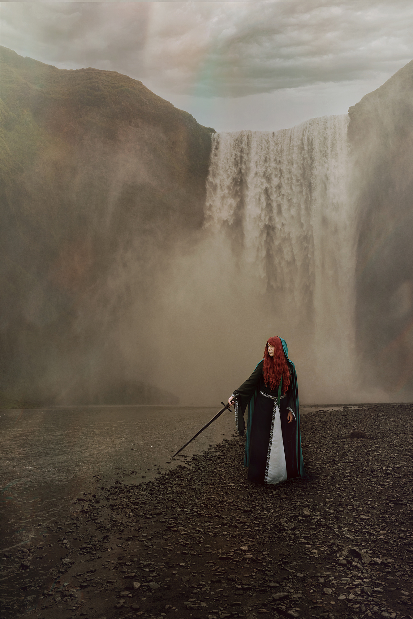 A white woman with red hair holds a sword and she poses in front of a waterfall wearing a fantastic costume consisting of a black and silver medieval dress and a green velvet cape. 