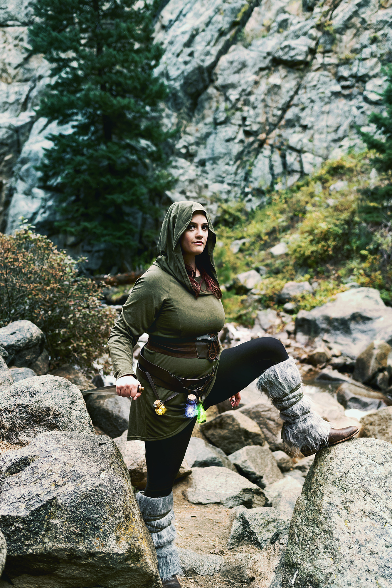 A white woman in a mage costume consisting of a green hooded tunic, black leggings, boots with gray fur, a dagger and a belt of magic potions poses on a rock in the woods for a fantasy photo by Kendra Colleen Photography.  