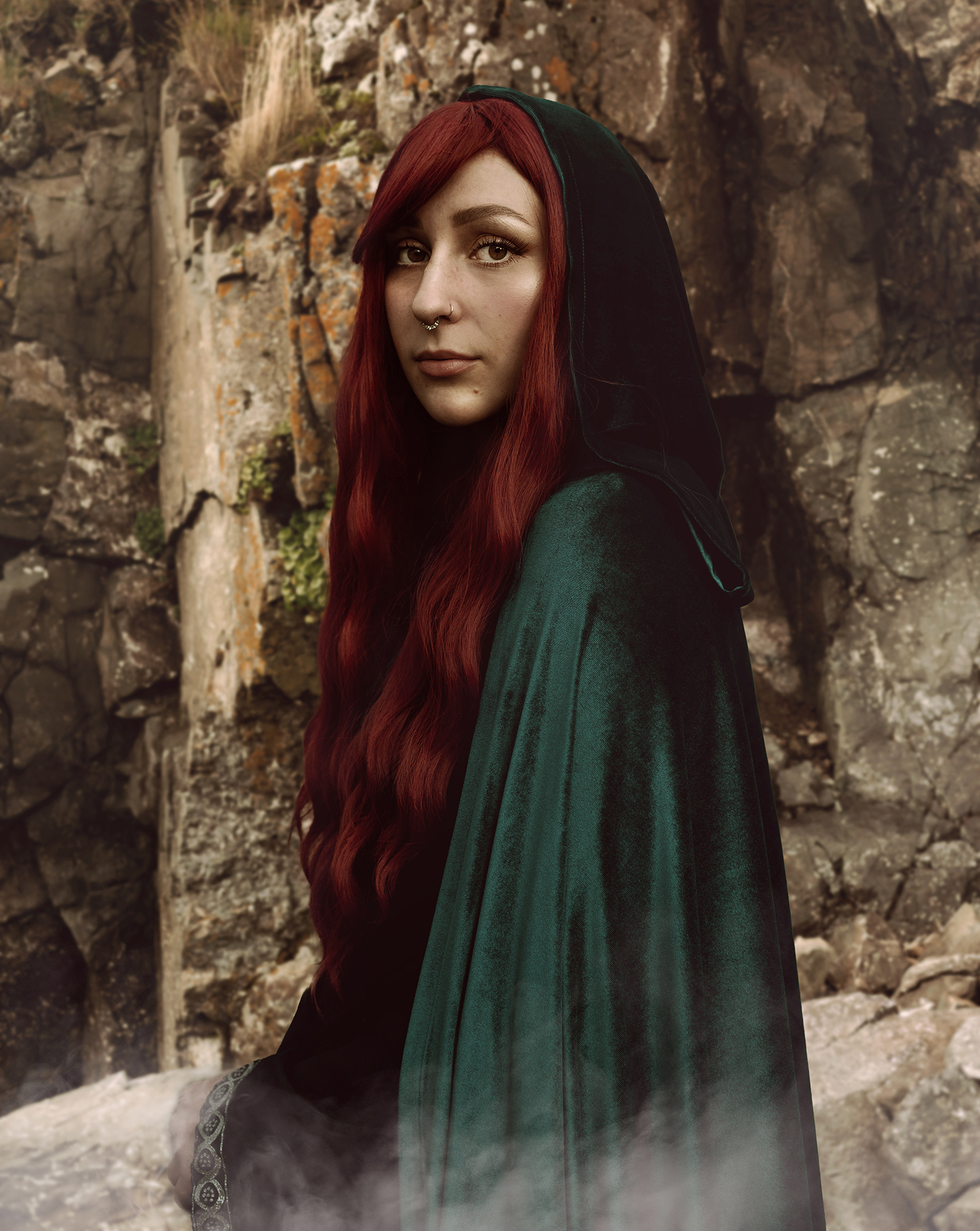 A white woman with dark red hair poses in front of a rock wearing a long velvet green cape for a fantasy photo by Kendra Colleen Photography in Longmont Colorado. 