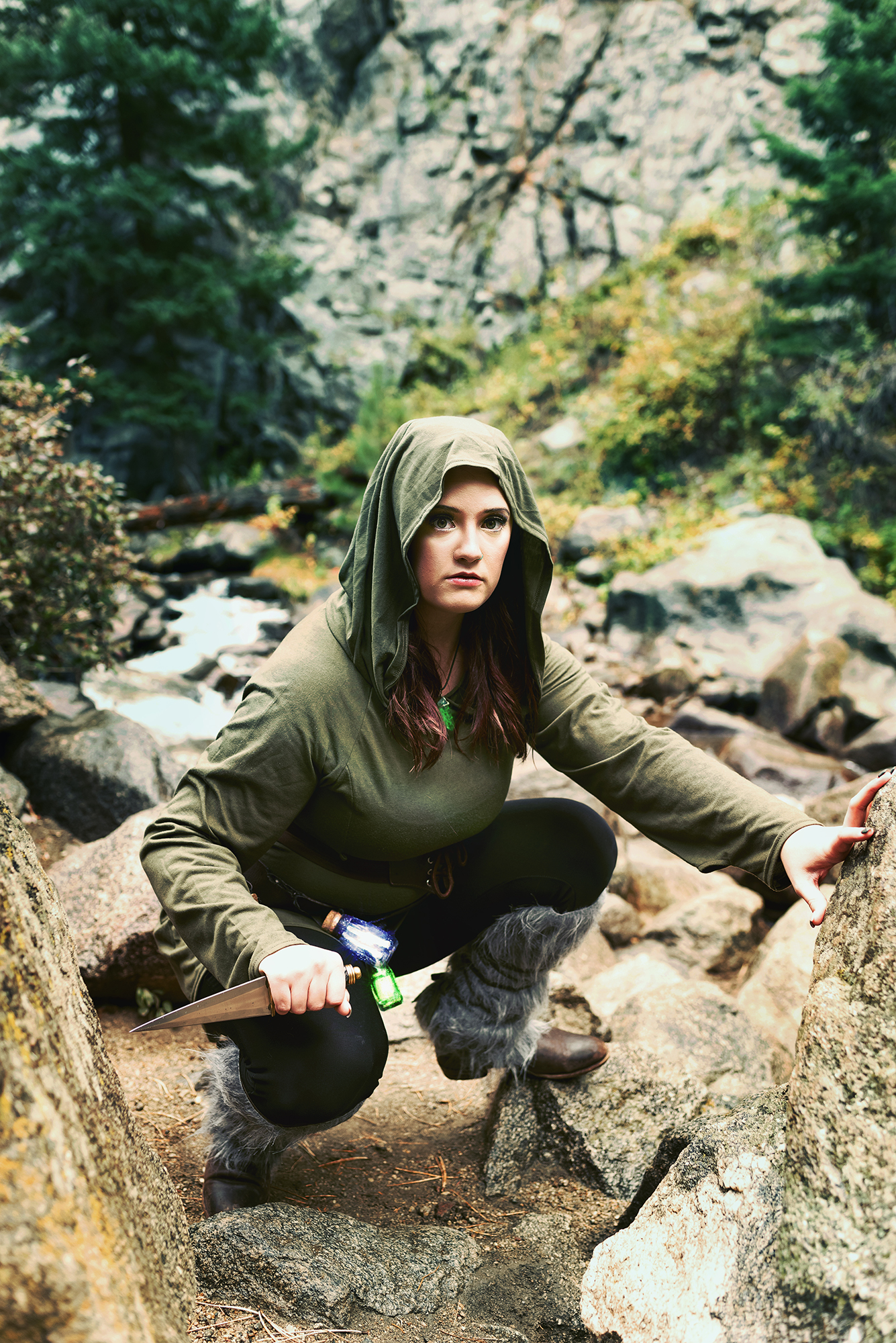 A white woman crouches between two rocks in the woods for a fantasy photo session, she holds a dagger in one hand and has magic potions strapped to her belt, she has dark hair and wears a green hooded tunic, black leggings and boots with gray fur.  