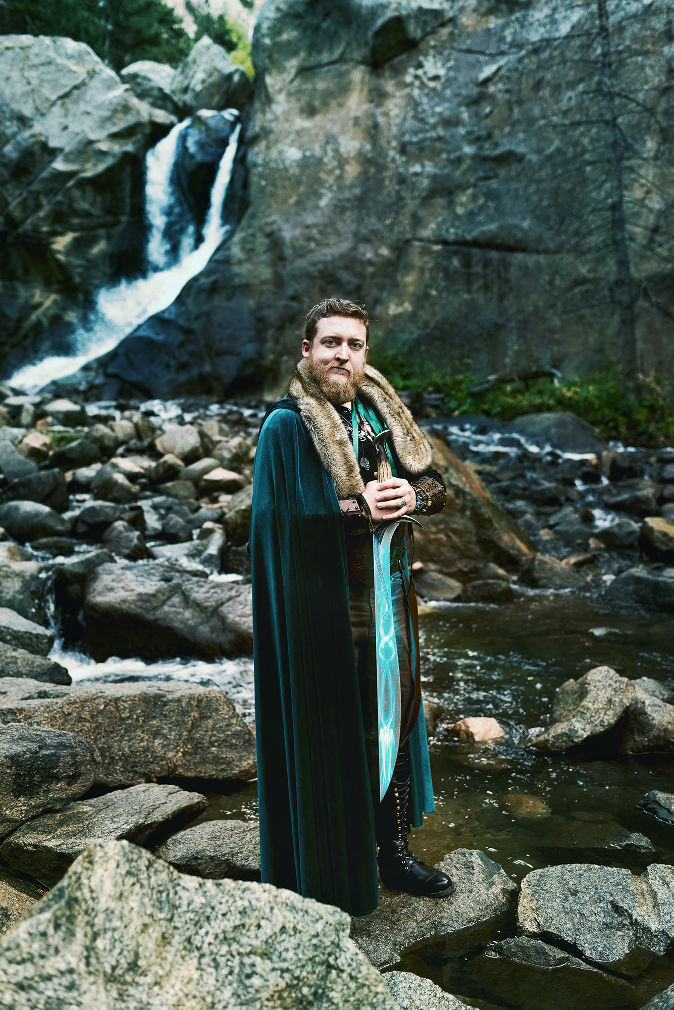 A white man in a Lord Of The Rings inspired costume consisting of a green velvet cloak with fur trim, a glowing sword and arm gauntlets poses on the rocks of a river for a fantasy photo session by Kendra Colleen Photography