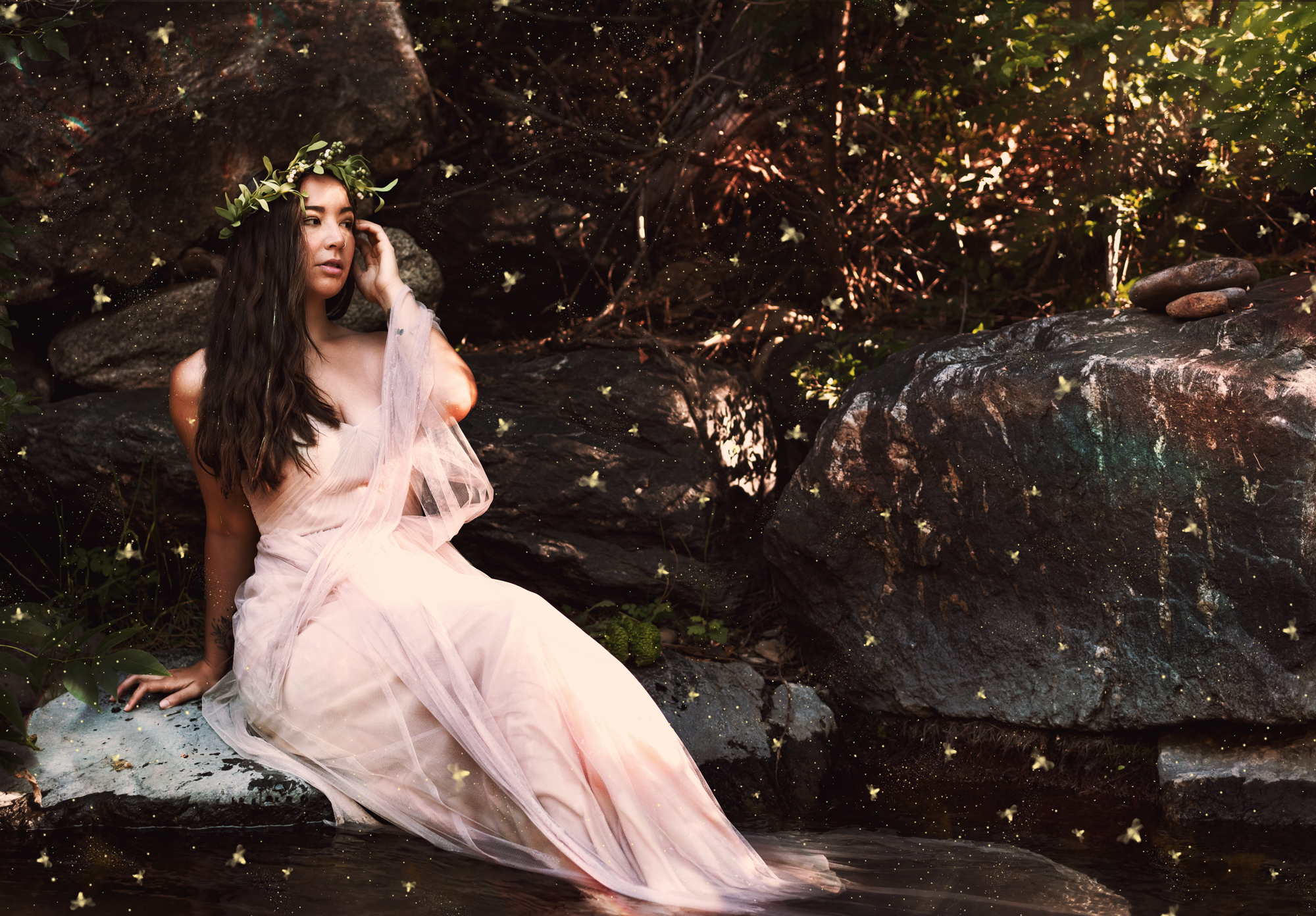 A fantasy photo of a brunette woman in a blush pink dress sitting on a rock with her feet in a creek with fireflies flying all around her. 