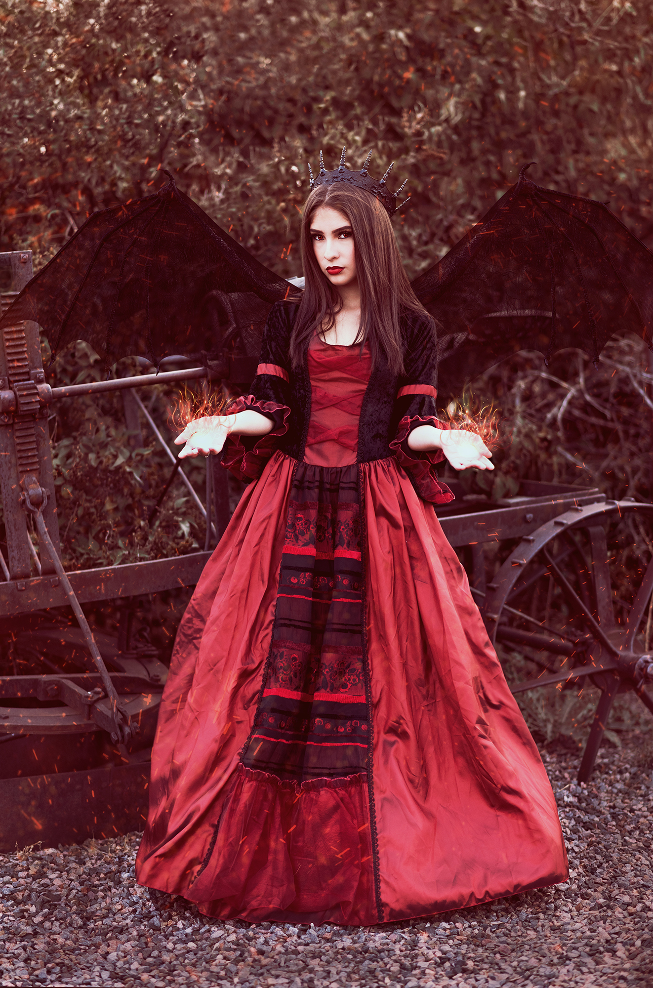 A fantasy photo by Kendra Colleen Photography of a girl dressed as a demon princess in a black and red gown who stands in front of old rusted equipment with black wings spread and flame magic in her outstretched hands.
