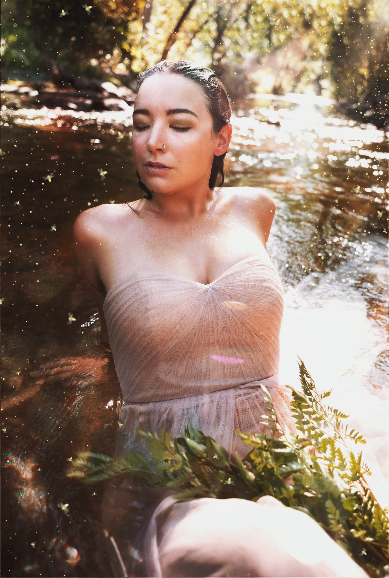 A fantasy photo by Kendra Colleen Photography of a woman laying in a shallow creek with magical fireflies and sparkles all around her in glittering sunlight. 
