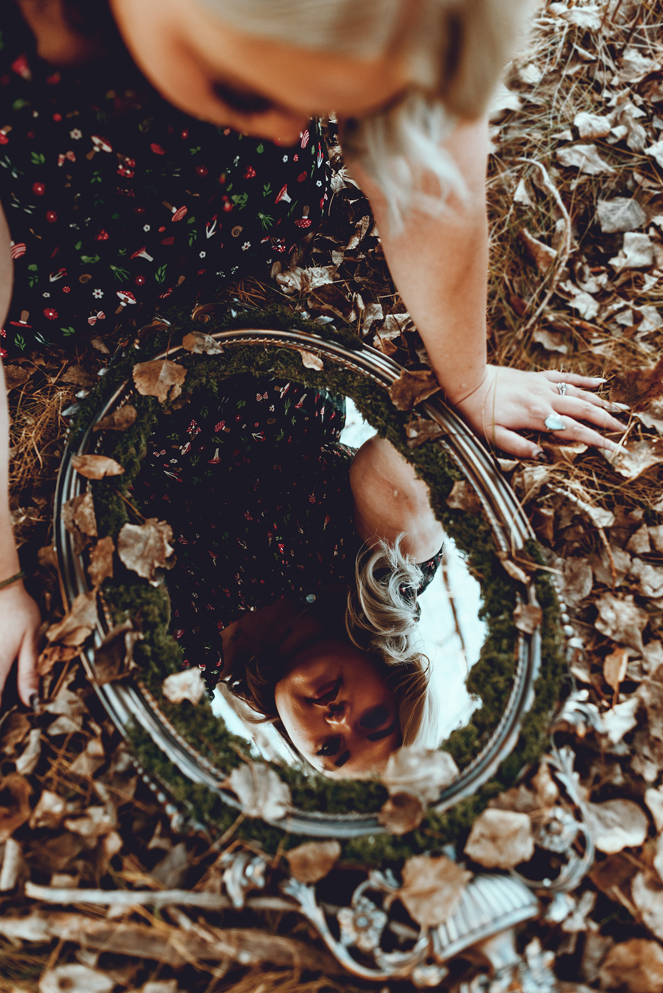 A person’s reflection in a round mirror on the ground, surrounded by dry leaves and twigs. The person is wearing a black dress with white flowers and their face is blurred to protect their privacy. Their hands are visible on either side of the mirror. The background consists of dry leaves and twigs