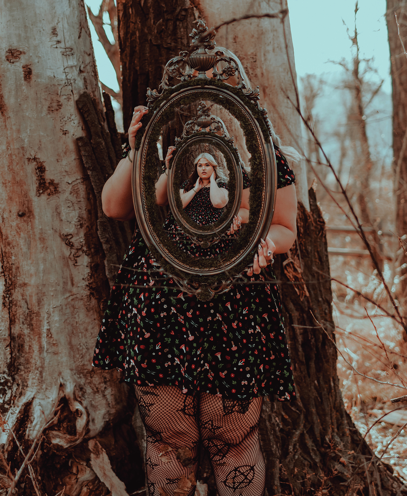 A person in a black floral dress stands behind a tree trunk holding an ornate gold mirror reflecting their face and upper body. The background is a forest with bare trees and dry leaves on the ground. The image has a warm, vintage tone