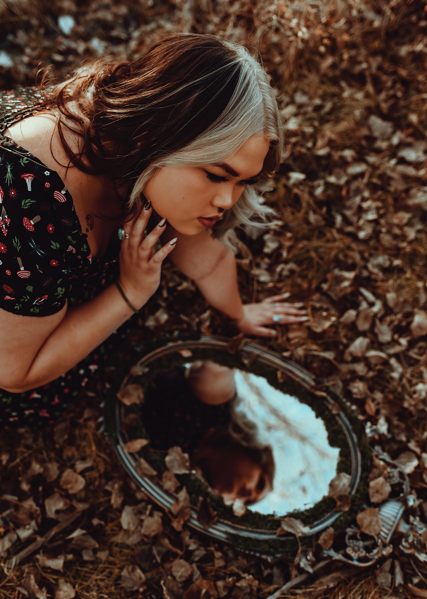 A person in a black floral dress kneels on the ground in a forest, looking at their blurred reflection in a round silver mirror. The ground is covered in fallen leaves and twigs, and the background consists of trees and foliage
