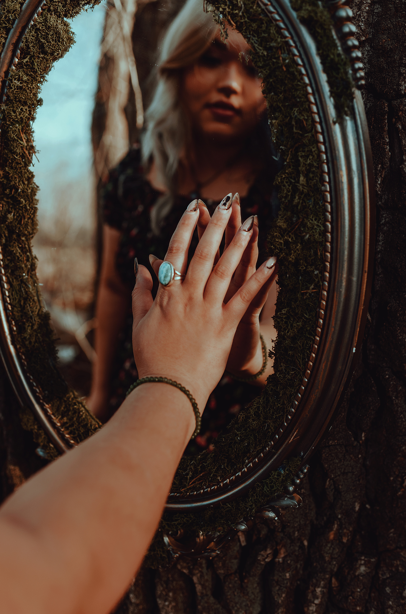 A person’s hands adorned with rings and bracelets in front of an oval gold-framed mirror attached to a tree trunk covered in moss. 