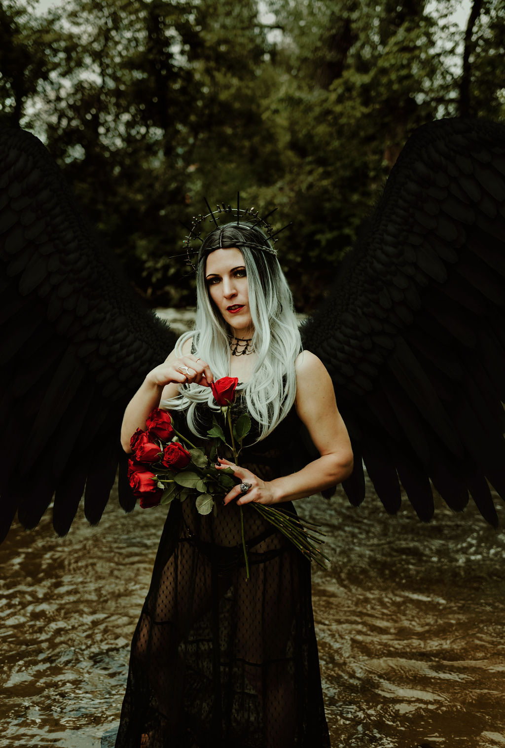 Kendra Colleen Photography takes fantasy image of a woman with large black wings holding a bouquet of red roses with a river and forest in the background.