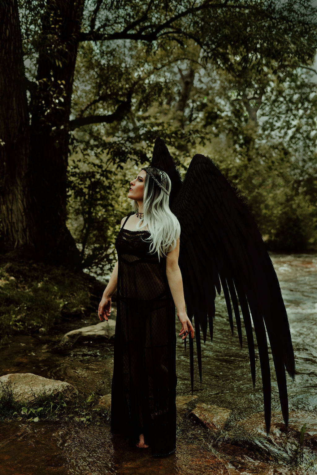 A woman stands in a forest with a river in the background. The woman has a pair of large black wings attached to her back