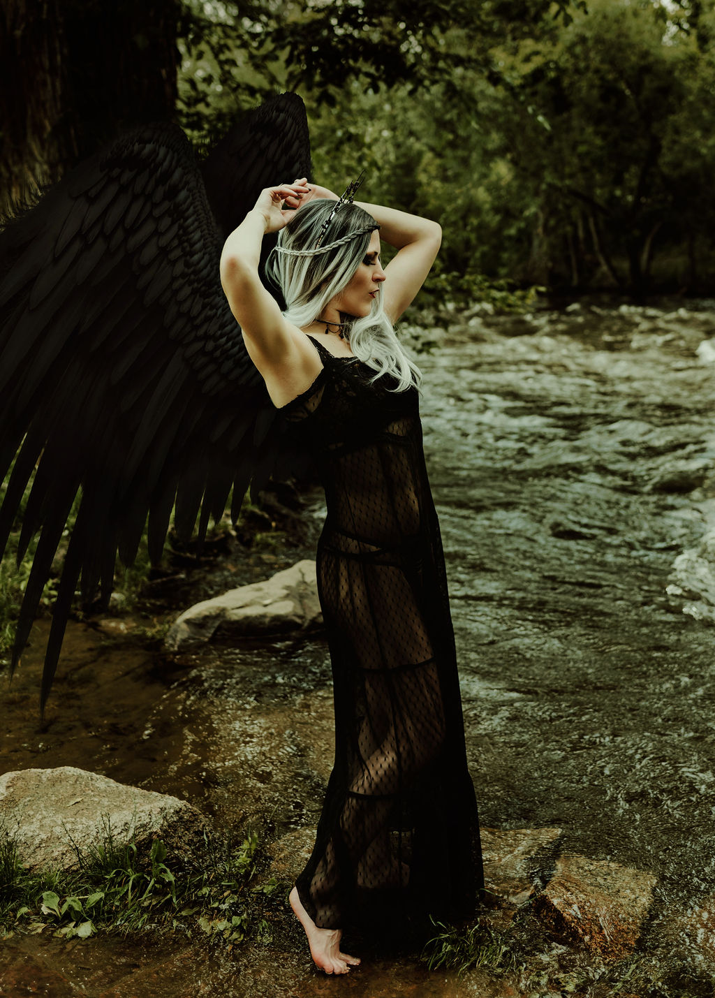 A woman stands in a forest with a river in the background. Her arms are raised above her head. The woman has a pair of large black wings attached to her back