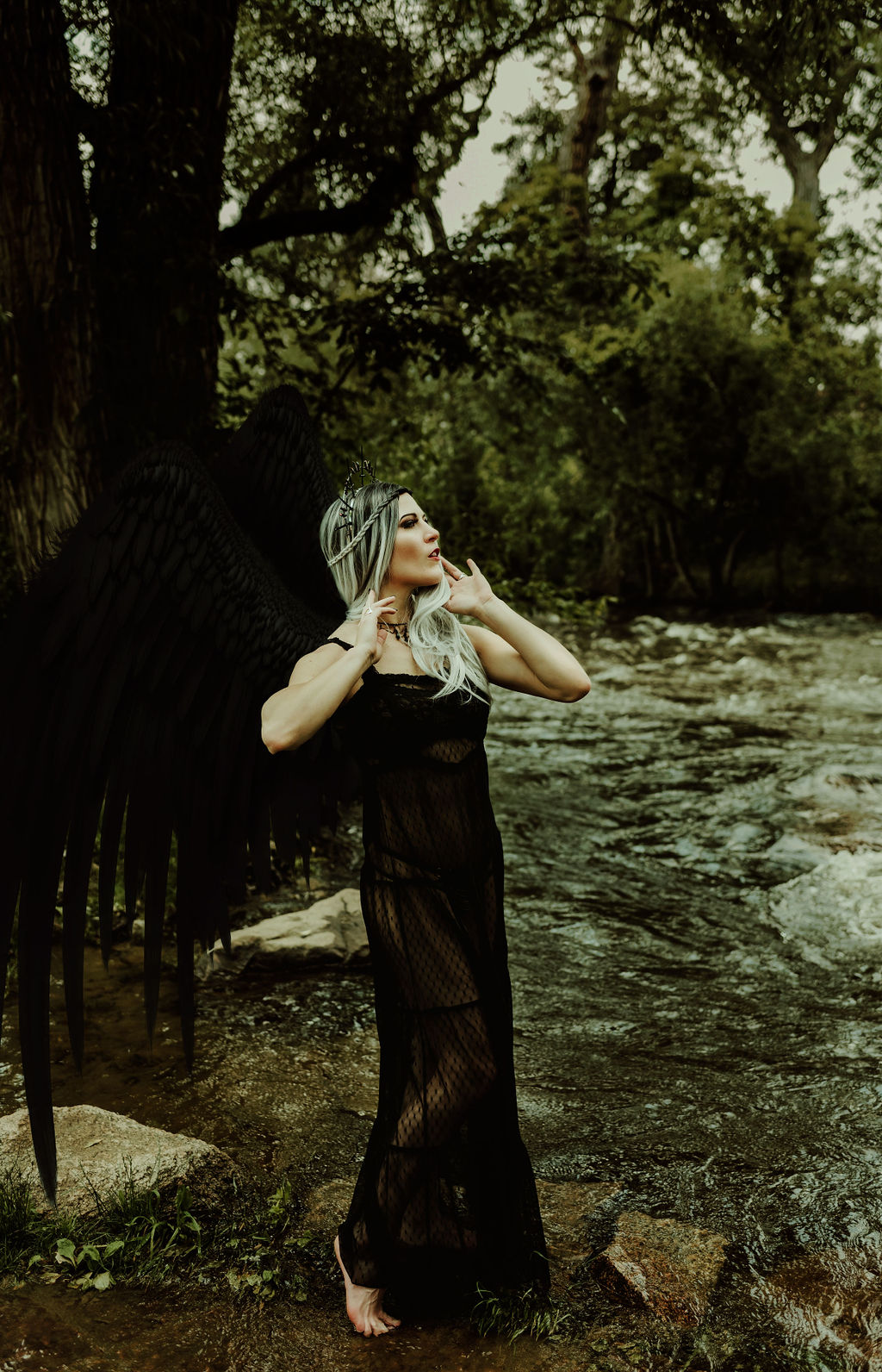 Kendra Colleen Photography takes fantasy boudoir photo of a woman in a black dress standing in a creek with a forest in the background. She has a pair of large black wings.
