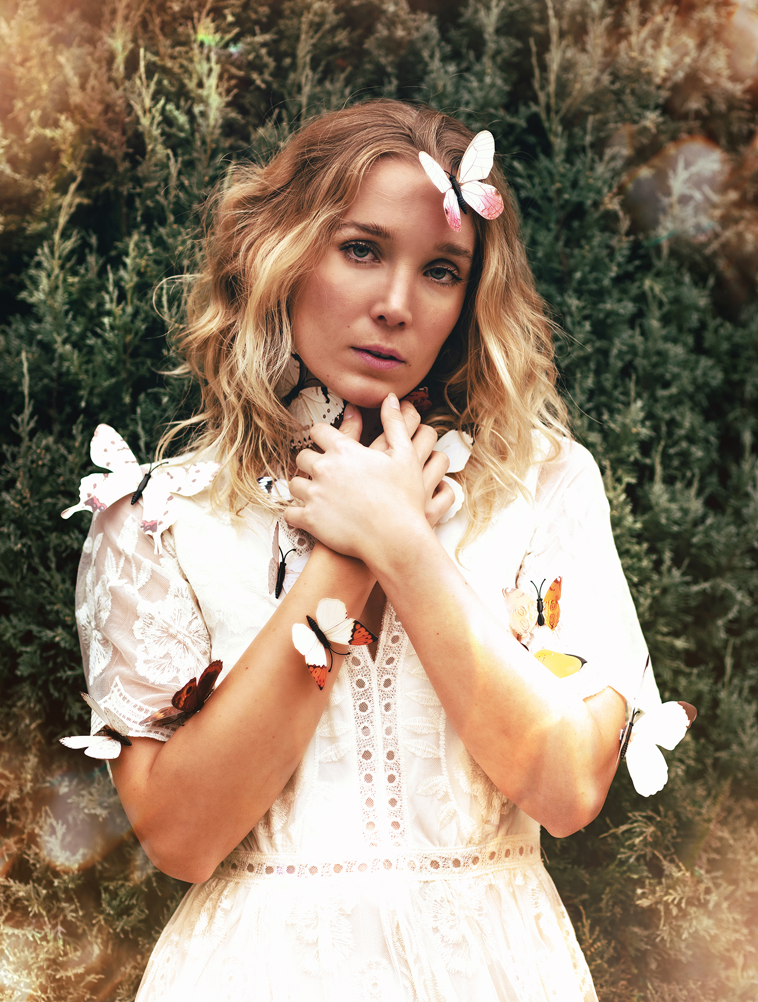 A person standing in front of a green bush, wearing a white dress with a lace collar and sleeves, and with orange and white butterflies stuck to their body.