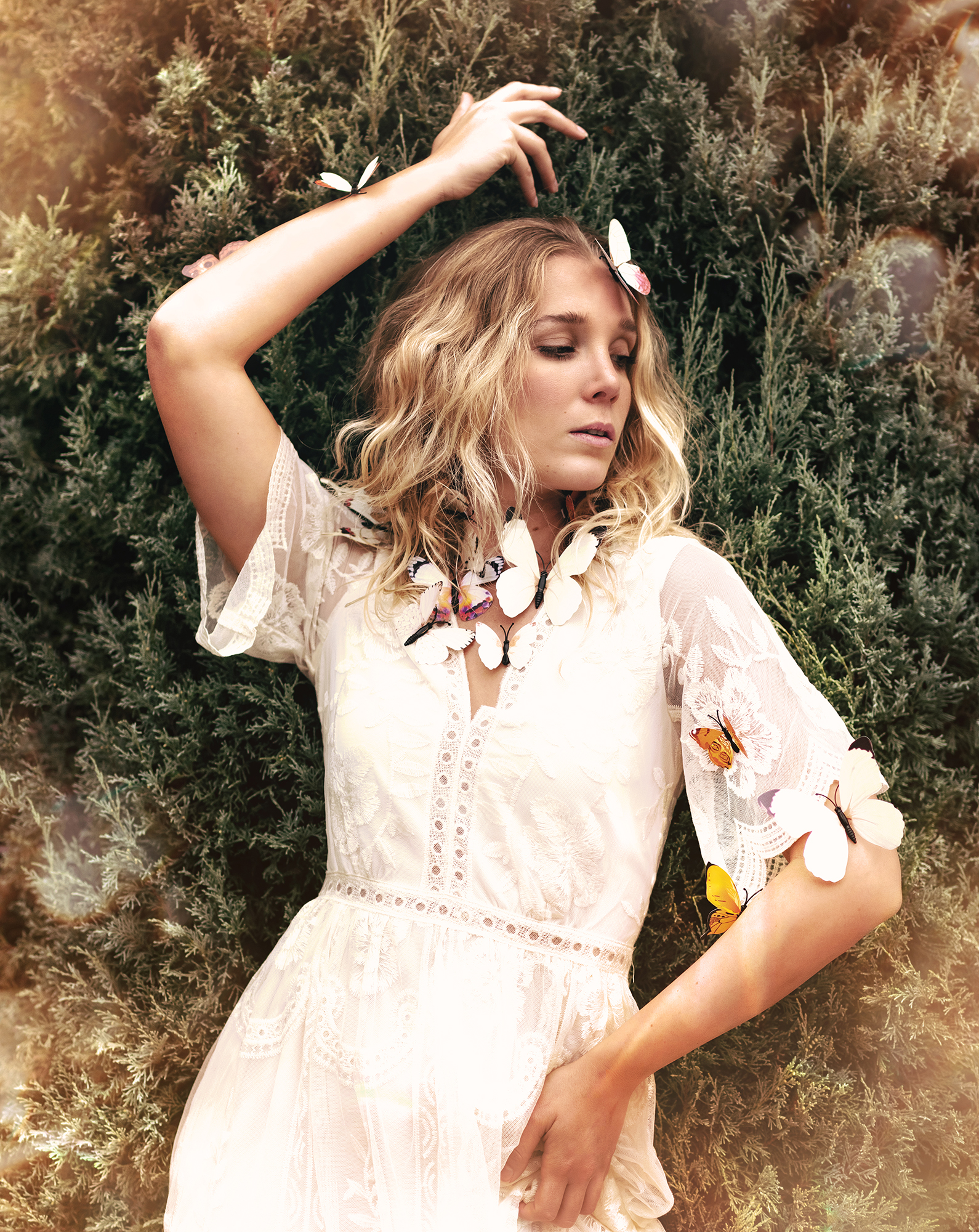A person standing in front of a greenery background with fake butterflies stuck to their body, wearing a white dress with a lace collar and sleeves