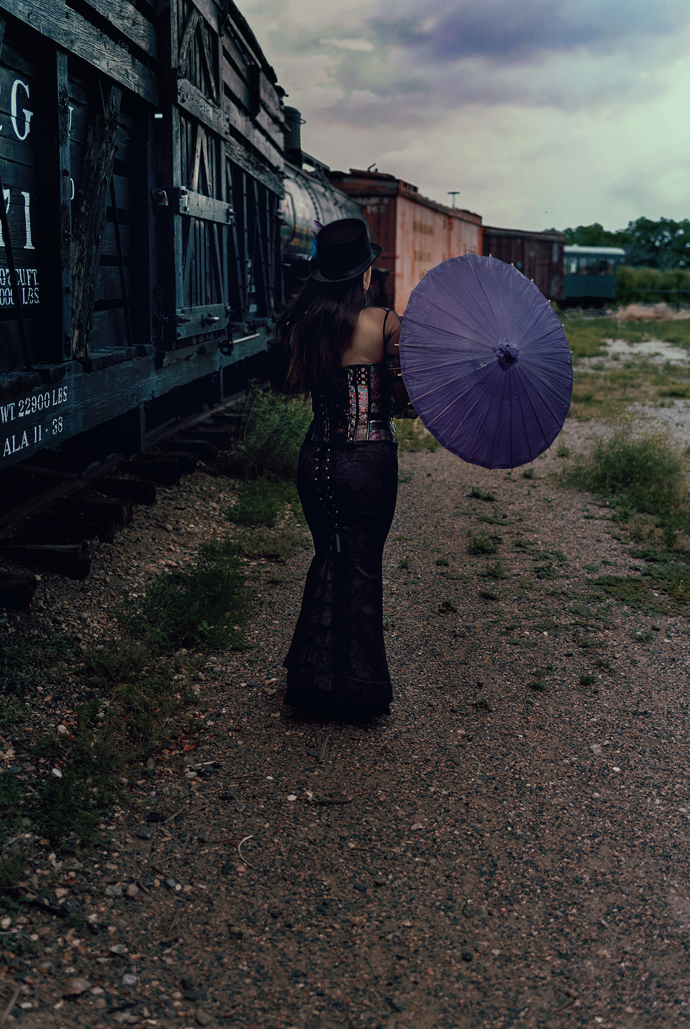 A photo of a woman from behind, she walks next to vintage train cars wearing a Victorian style steampunk outfit and holding a purple parasol.