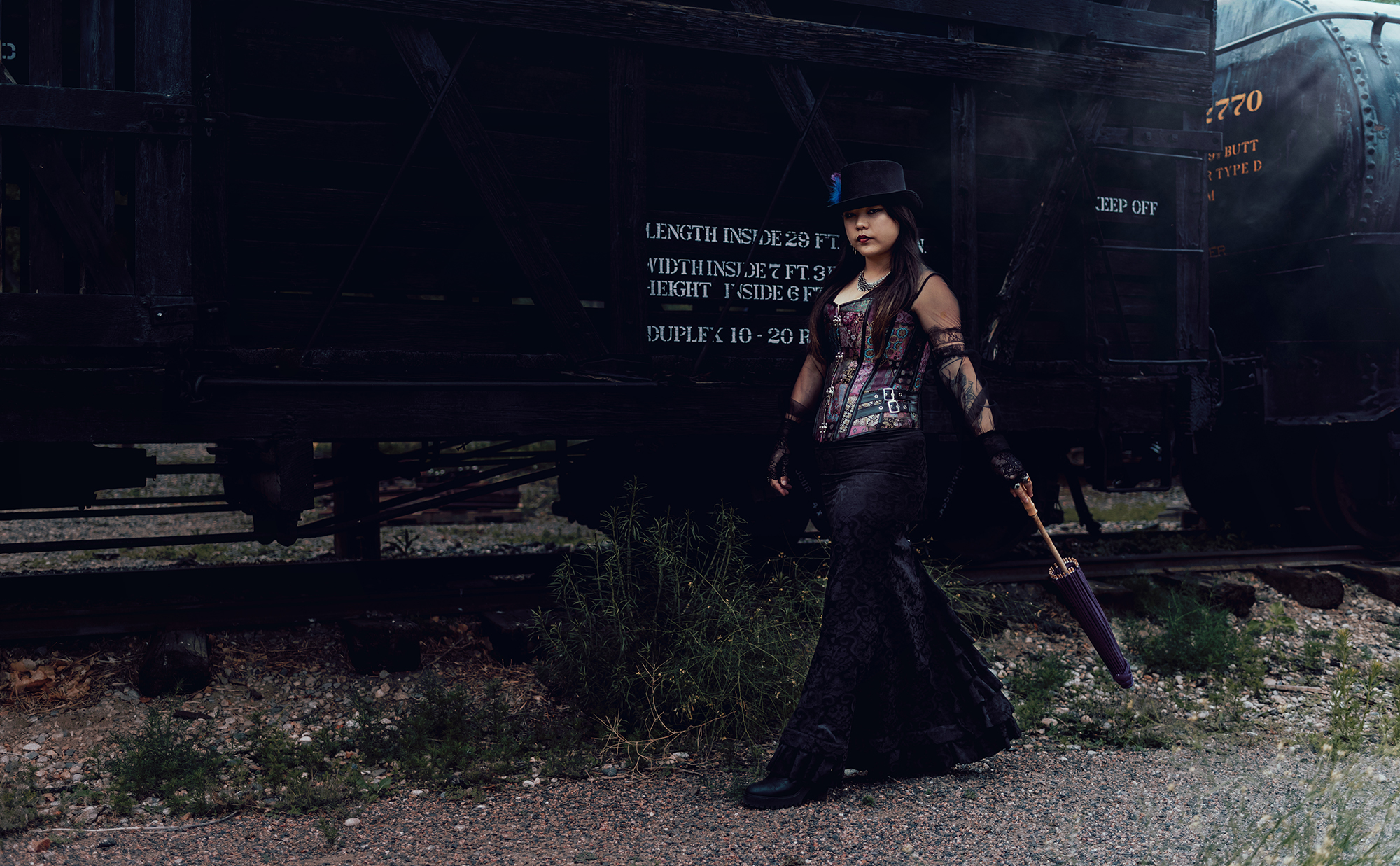 A woman walks next to a wooden train car wearing a Victorian style steampunk outfit holding a purple parasol