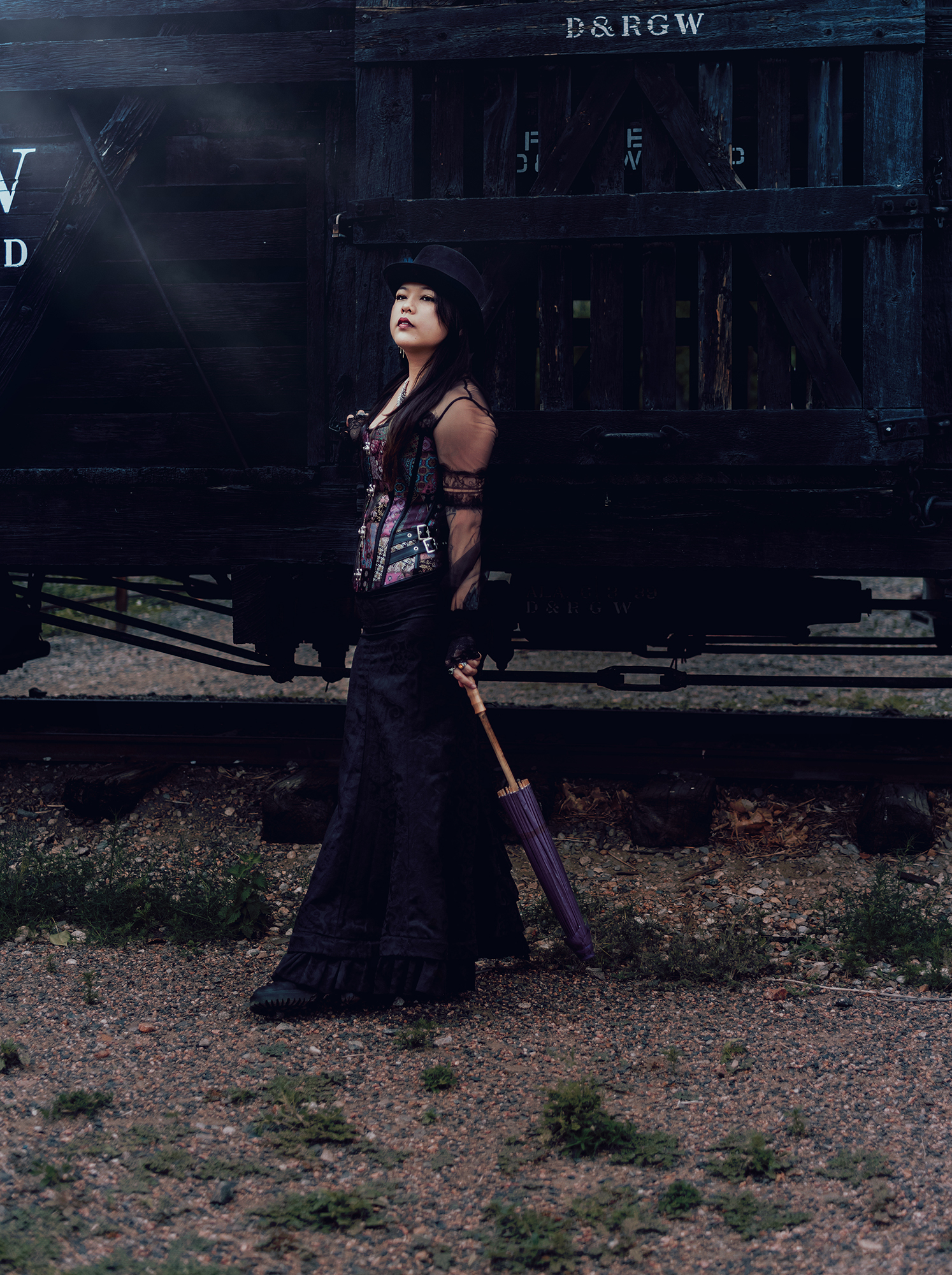A woman standing next to a wooden train car wearing a Victorian style steampunk outfit and holding a purple parasol
