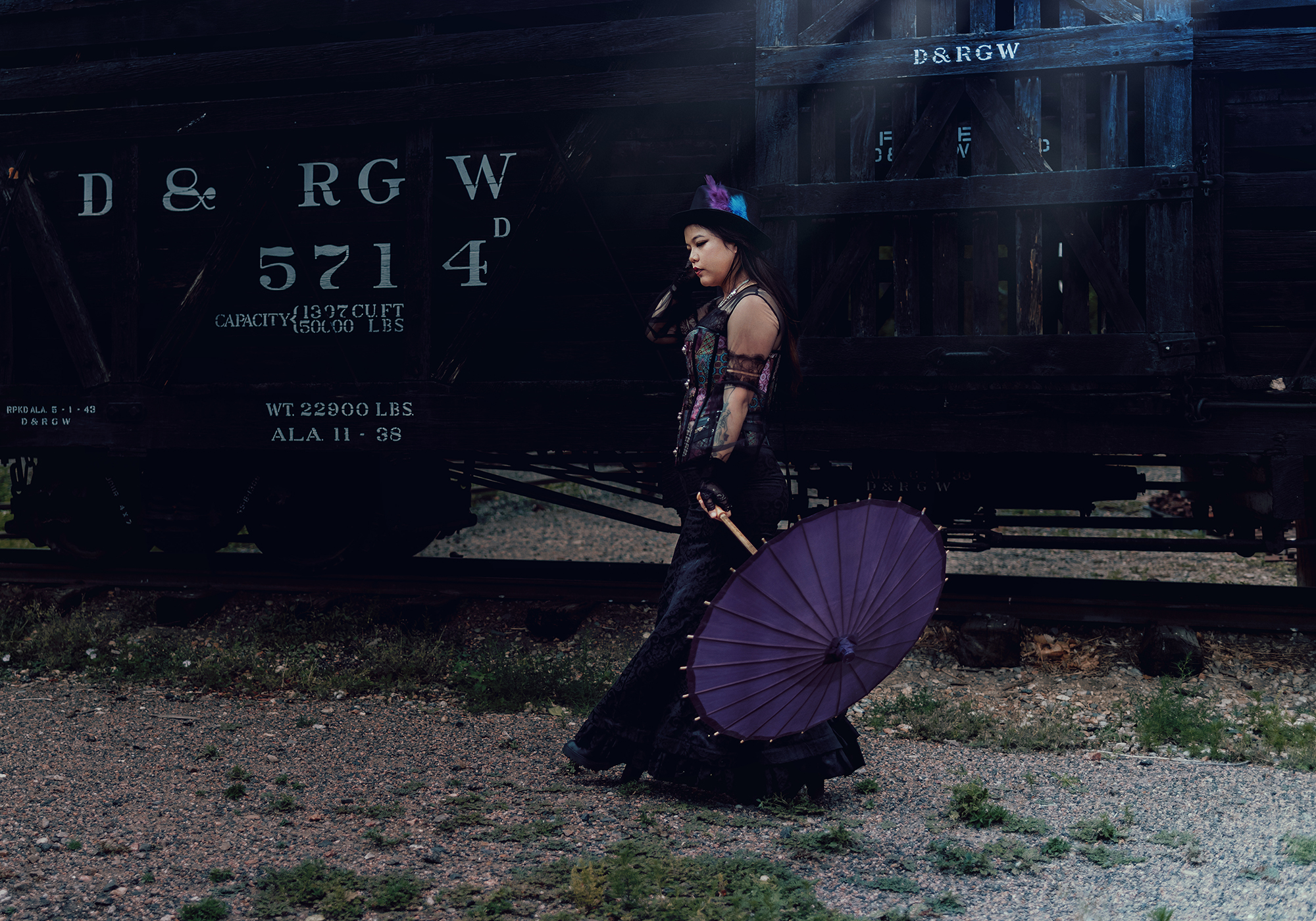 A fantasy photo of a woman walking next to a wooden train car in a Victorian style steampunk outfit holding a purple parasol