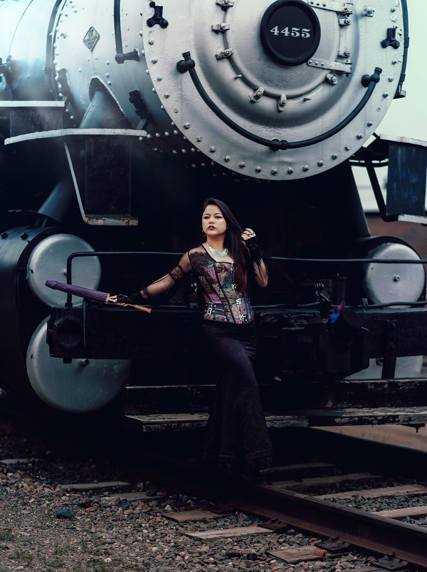 A steam engine train with a woman standing in front of it wearing a Victorian style steampunk outfit for fine art portrait by Kendra Colleen Photography