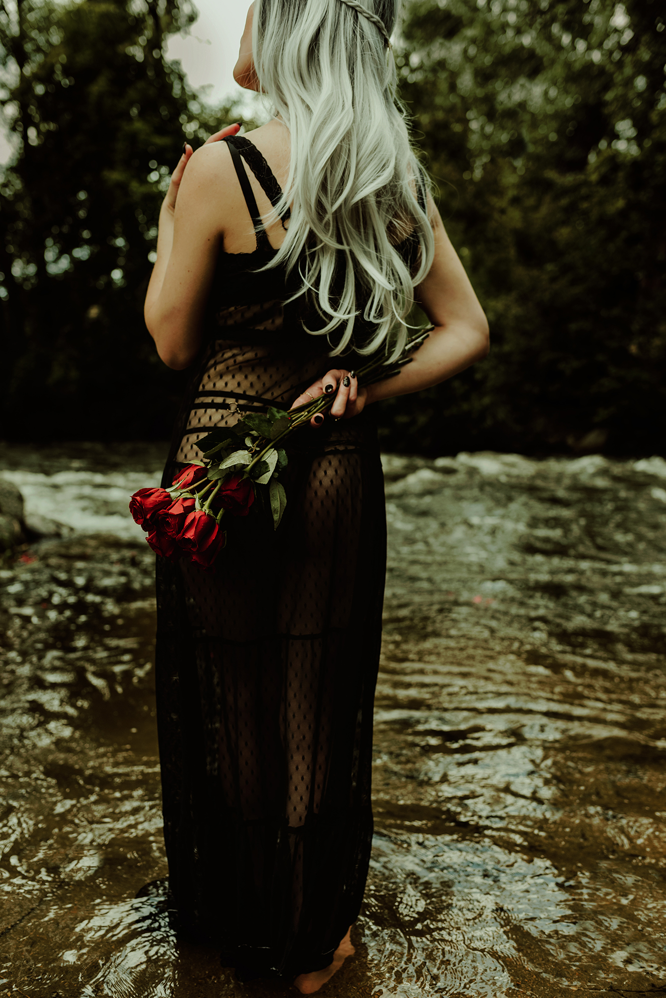 A white woman with silver hair, wearing a sheer black dress, viewed from the back, holds a bouquet of roses behind her back for boudoir portrait by Kendra Colleen Photography