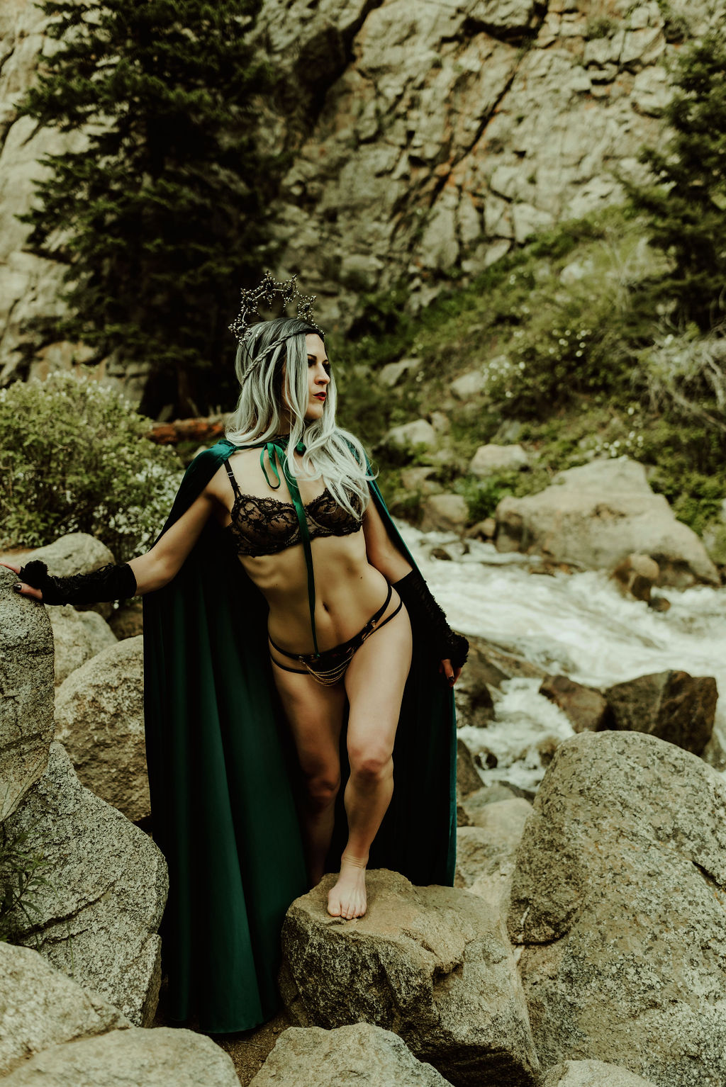 A white woman with silver and black hair poses in front of a waterfall wearing black lingerie, a dark green cape and a goth headpiece for a boudoir photo by Kendra Colleen Photography