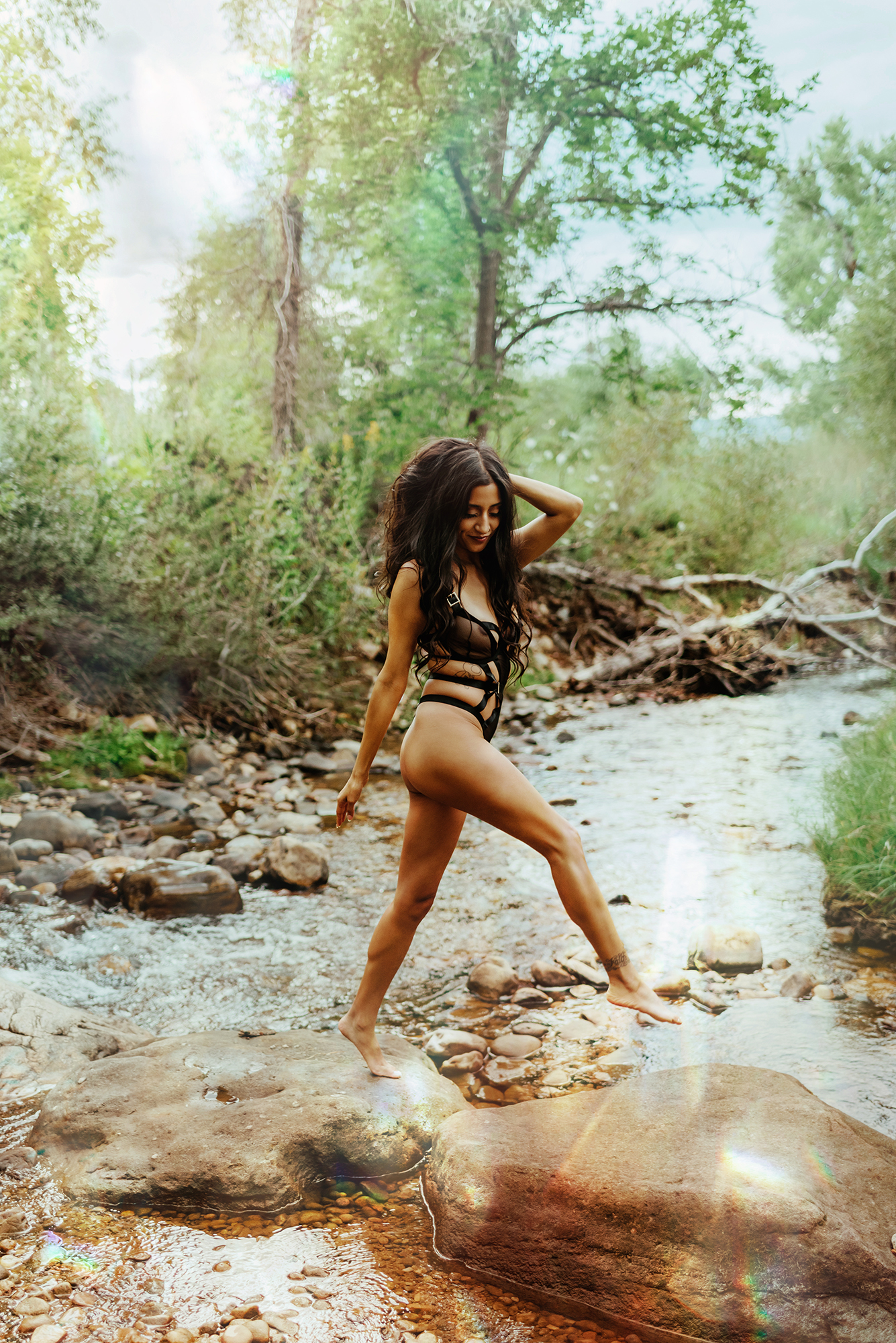 A Latina girl in black, strappy lingerie jumps from one rock to another in a creek surrounded by trees for Boudoir photos in Boulder Colorado. 